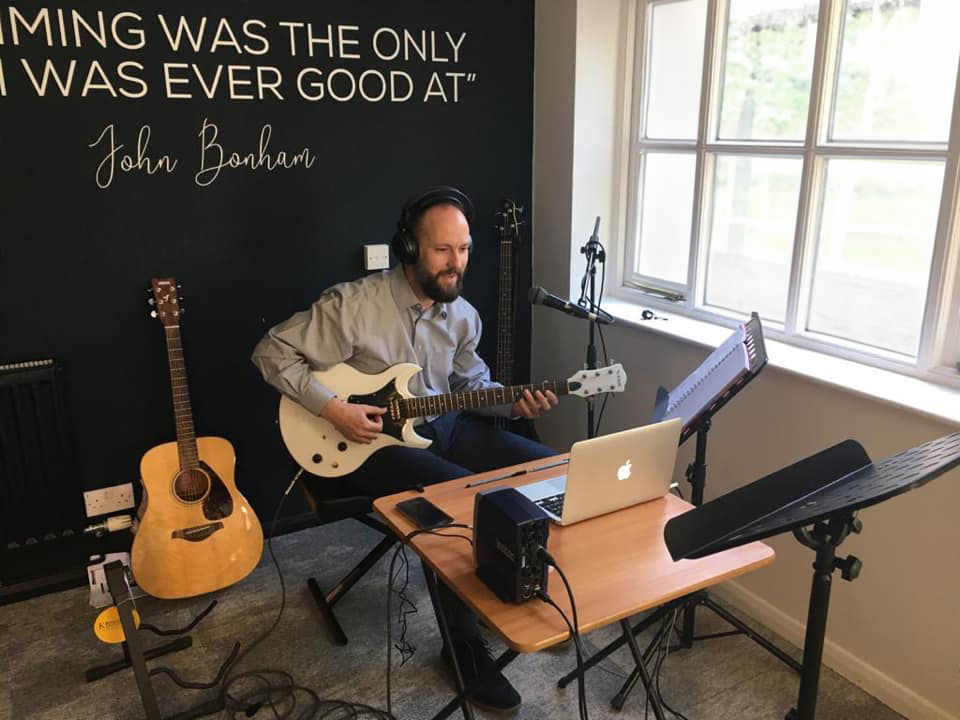 HCM tutor doing an online lesson with a guitar in hand and a laptop in front of him.