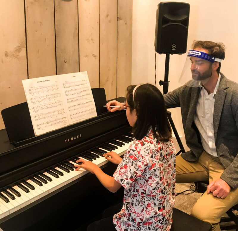 Tutor teaching piano to a girl whilst wearing PPE